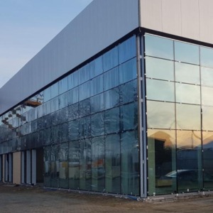 Elewacja budynku biurowego z dużymi, szklanymi panelami okiennymi, odbijającymi zachodzące słońce i krajobraz. Widoczna konstrukcja budynku i fragment placu budowy.