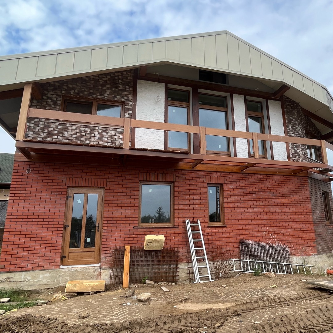 Ceglany dom z balkonem, widoczne okna z drewnianą ramą, drzwi wejściowe i drabina oparta o ścianę, teren wokół domu w trakcie prac wykończeniowych.
