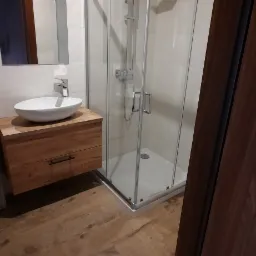 Nowoczesna łazienka z kabiną prysznicową, umywalką nablatową na drewnianej szafce i podłogą z paneli imitujących drewno ułożonych w jodełkę.