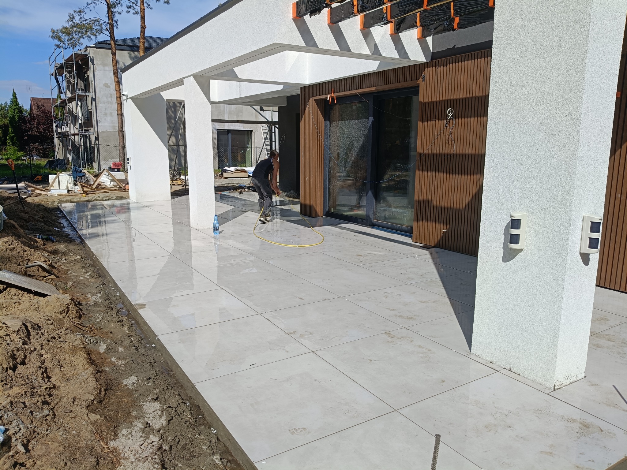Białe, błyszczące płytki tarasowe ułożone przed nowoczesnym budynkiem. Pracownik czyści powierzchnię. Widoczne narzędzia i elementy konstrukcyjne.