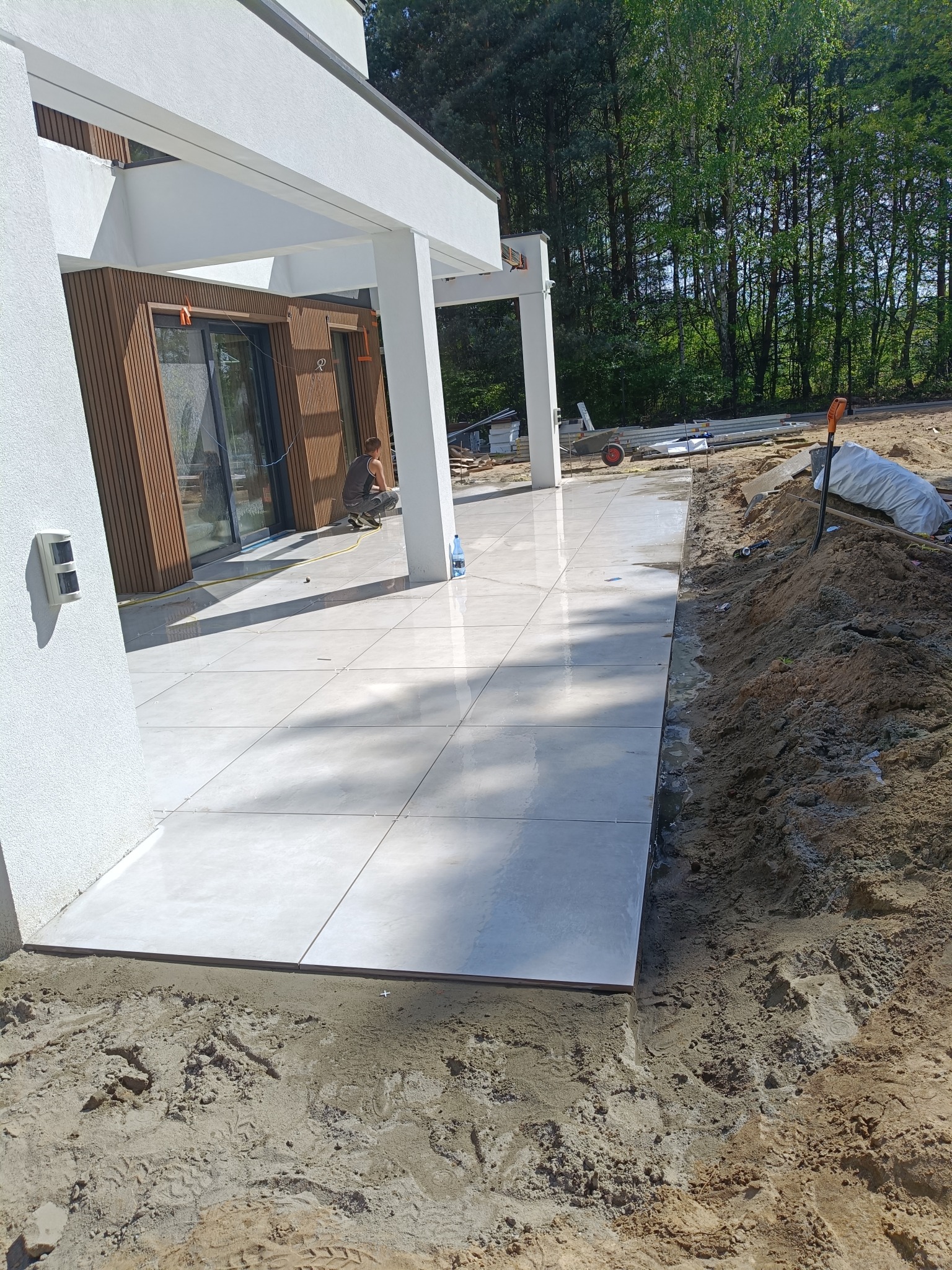 Jasne płytki tarasowe ułożone przy nowoczesnym domu z drewnianą elewacją. W tle widoczny pracownik podczas fugowania i narzędzia.