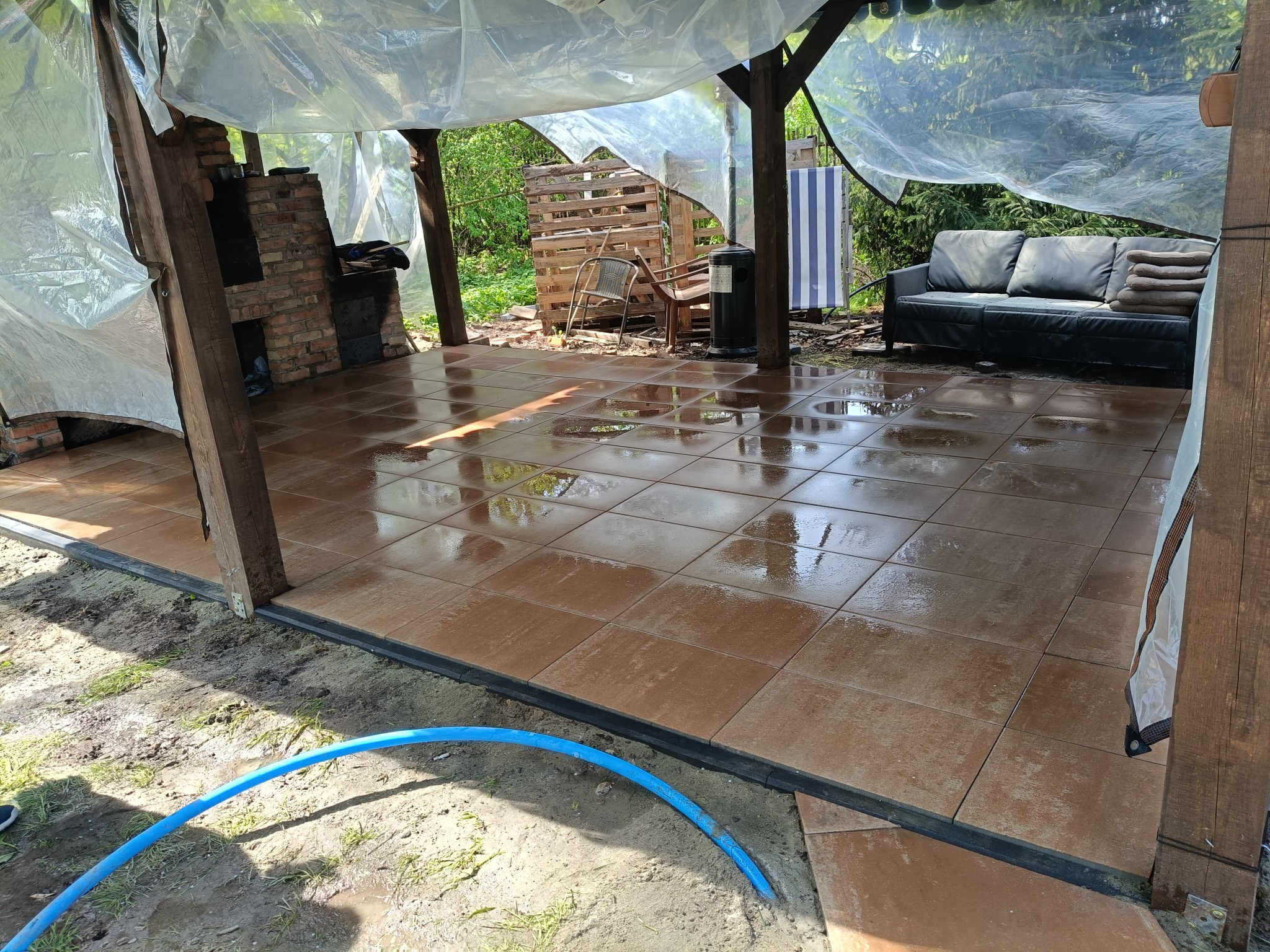 Ułożona, brązowa terakota na zewnątrz z meblami ogrodowymi pod zadaszeniem. Widoczny fragment niezagospodarowanego terenu z niebieskim wężem ogrodowym.