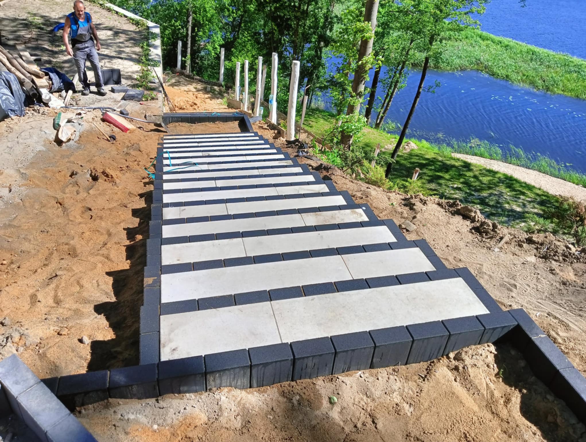 Schody z kostki brukowej w trakcie budowy, z widokiem na jezioro i pracownika w tle. Kostka w kolorach szarym i czarnym, ułożona na zboczu.