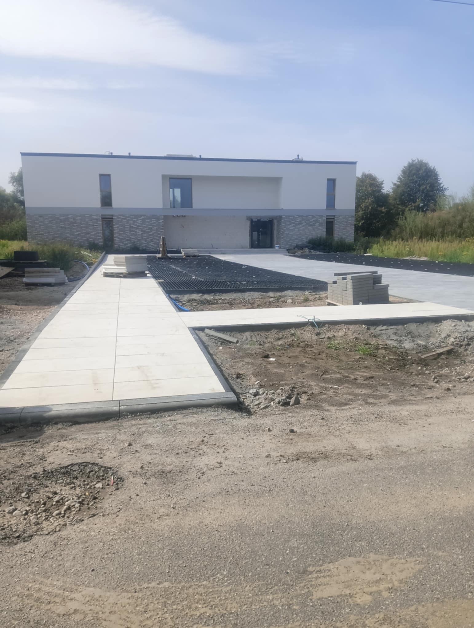 Jasne płyty chodnikowe i podjazd z ażurowych płyt na posesji przed domem. Częściowo ułożona kostka brukowa, widoczne przygotowania do dalszych prac.