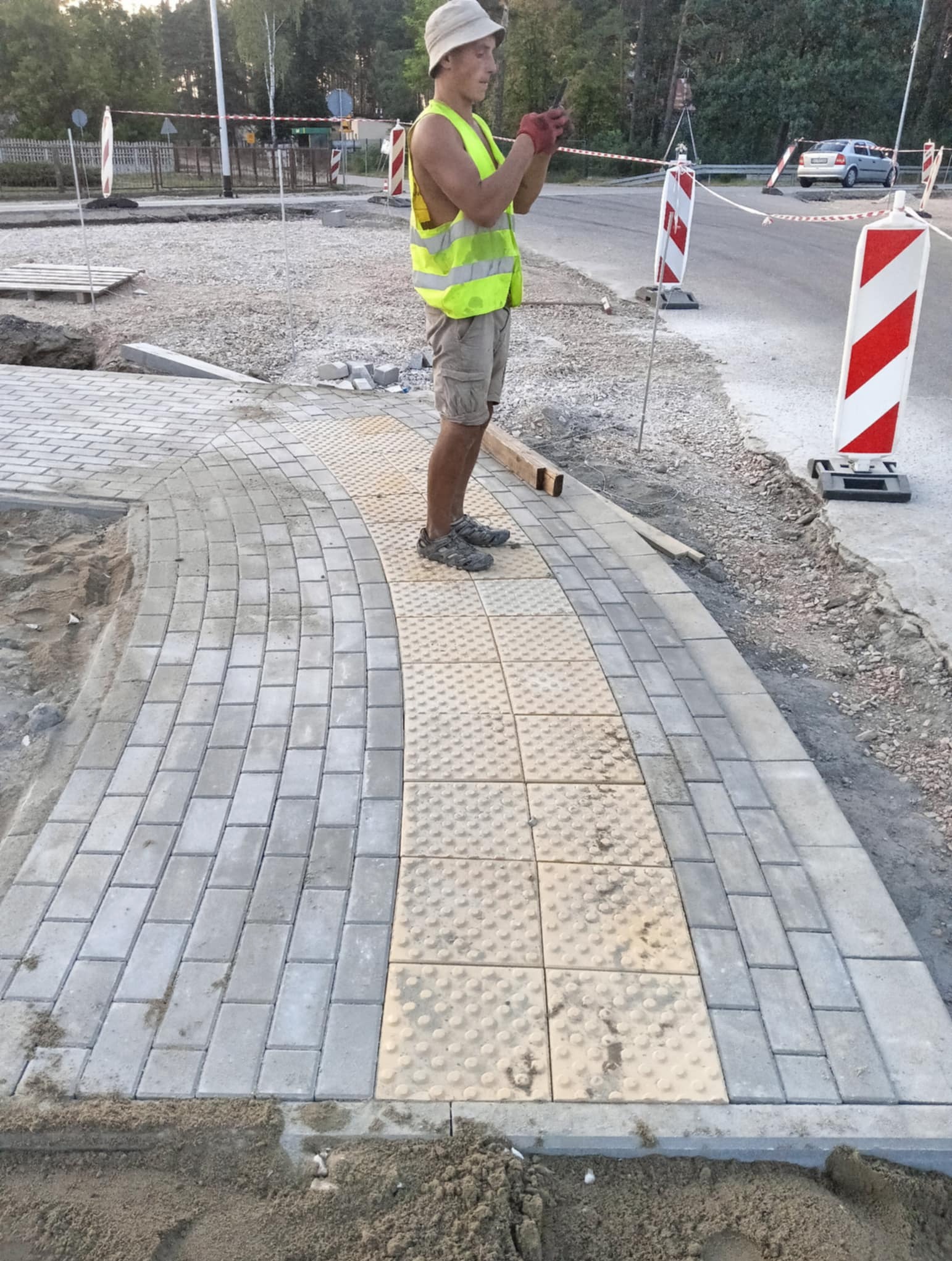 Nowa ścieżka z kostki brukowej z elementami dla niewidomych, wykonawca w kamizelce odblaskowej i kapeluszu, w tle droga i znaki drogowe.