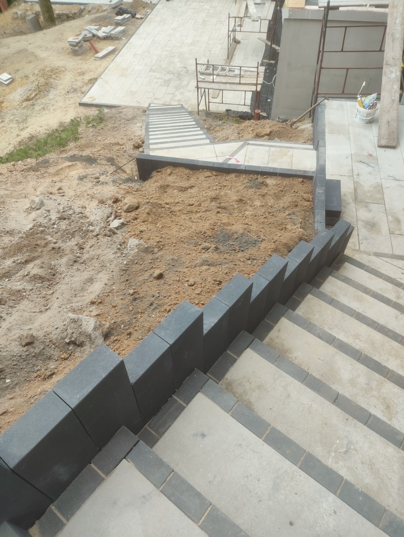 Schody z kostki brukowej w trakcie budowy, widok z góry. Część terenu nieutwardzona. Widoczne materiały budowlane i rusztowania w tle.