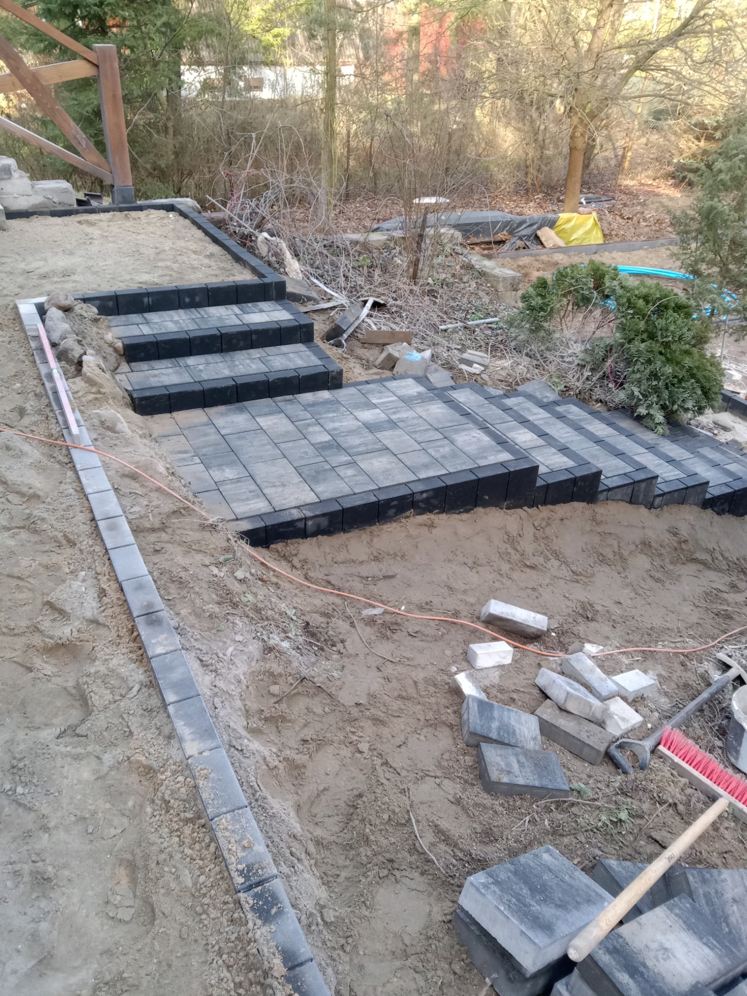 Schody z kostki brukowej w trakcie budowy, widoczne narzędzia i materiały, w tle drzewa. Czarno-szara kostka tworzy stopnie na nierównym terenie.