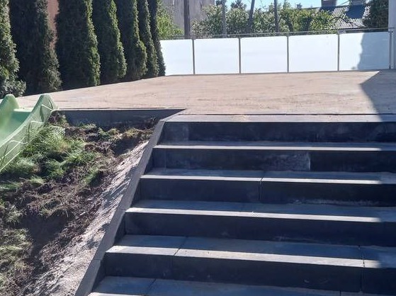 Schody z kostki brukowej prowadzące na taras z balustradą, obok zjeżdżalnia. Widoczny fragment ogrodu z tujami. Nowoczesne wykończenie przestrzeni.