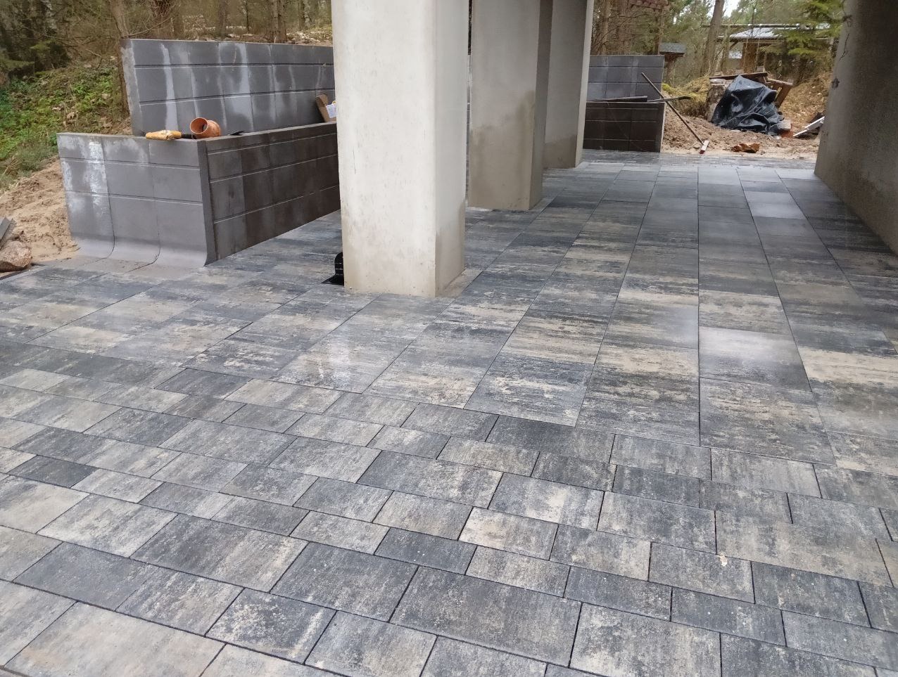 Ułożona szara kostka brukowa na podwórzu, z betonowymi filarami i elementami architektury ogrodowej w tle. Widok z perspektywy żabiej.