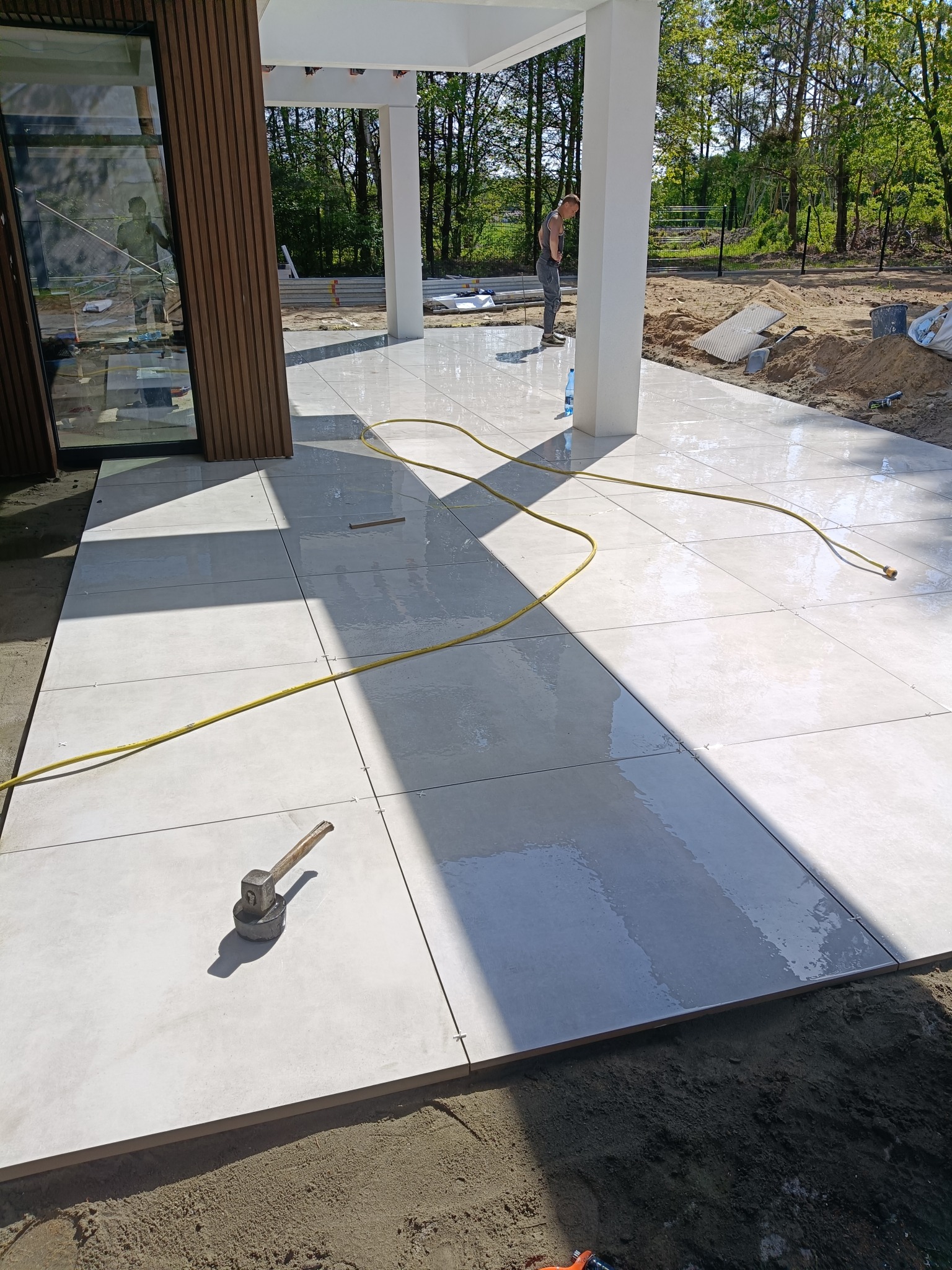 Jasne, kwadratowe płyty ułożone na tarasie, widoczny młotek gumowy, żółty wąż i pracownik w tle. Etap prac wykończeniowych z zachowaniem odstępów między płytami.