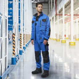 Mężczyzna w niebieskim kombinezonie roboczym z czarnymi wstawkami, stojący w hali magazynowej z regałami.