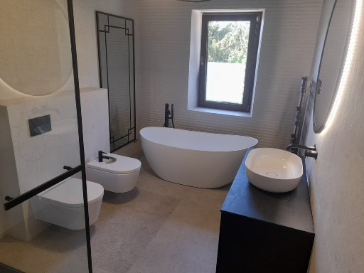 Nowoczesna łazienka z wolnostojącą wanną, podwieszaną toaletą i bidetem, czarną armaturą oraz dużym oknem z widokiem na zieleń.