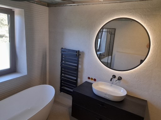 Nowoczesna łazienka z owalną umywalką nablatową, okrągłym lustrem z podświetleniem LED, grafitowym grzejnikiem drabinkowym i wolnostojącą białą wanną przy oknie.