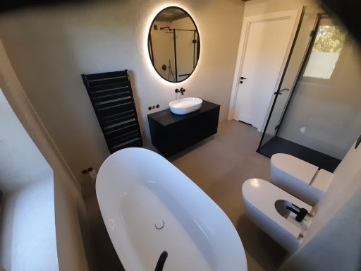 Nowoczesna łazienka z wolnostojącą wanną, podświetlanym okrągłym lustrem, czarną szafką pod umywalką i grzejnikiem drabinkowym w ciemnym kolorze, oraz kabiną prysznicową typu walk-in z czarnymi...