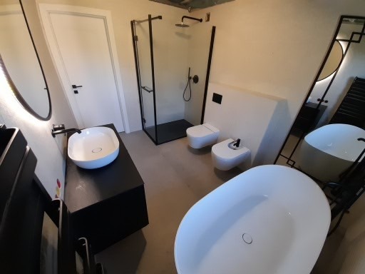 Nowoczesna łazienka z wolnostojącą białą wanną, umywalką nablatową na czarnym blacie, kabiną prysznicową typu walk-in z czarnymi profilami oraz toaletą i bidetem w minimalistycznym stylu.