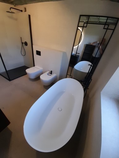 Nowoczesna łazienka z wolnostojącą wanną, toaletą i bidetem w białej kolorystyce, czarną kabiną prysznicową typu walk-in oraz lustrem w czarnej ramie o geometrycznym wzorze.