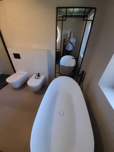 Nowoczesna łazienka na poddaszu z białą wanną wolnostojącą, toaletą i bidetem w minimalistycznym stylu. W lustrze odbija się osoba robiąca zdjęcie.