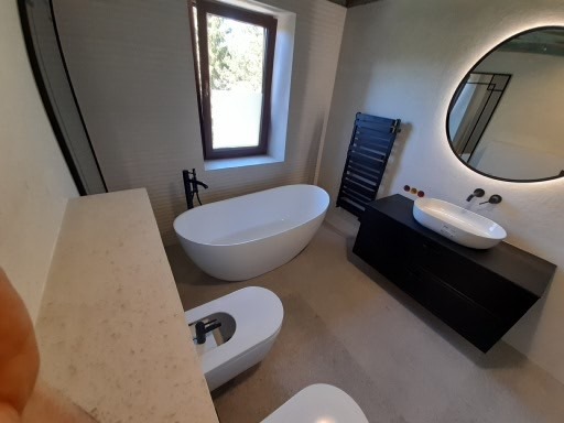 Nowoczesna łazienka z wolnostojącą wanną, owalnym lustrem z podświetleniem LED, wiszącą szafką z umywalką oraz grzejnikiem drabinkowym w ciemnym kolorze.