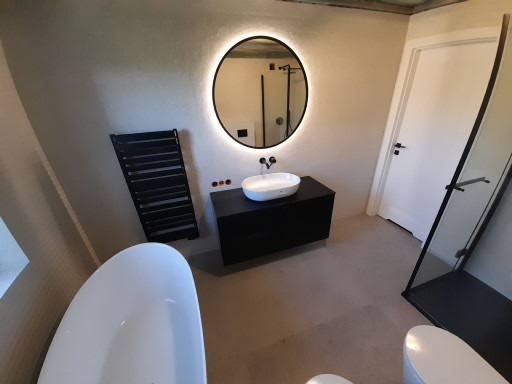 Nowoczesna łazienka z owalnym lustrem podświetlanym LED, czarną szafką pod umywalką nablatową i grzejnikiem drabinkowym, kabina prysznicowa typu walk-in z czarnymi profilami.