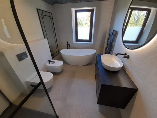 Nowoczesna łazienka z wolnostojącą wanną, umywalką nablatową na ciemnej szafce, wiszącą toaletą i bidetem, oraz podświetlanym lustrem owalnym.