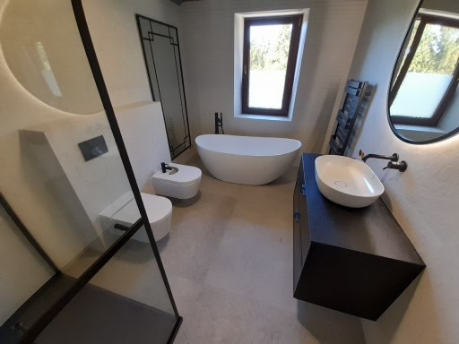 Nowoczesna łazienka z wolnostojącą wanną, umywalką nablatową na ciemnej szafce, toaletą i bidetem w minimalistycznym stylu, z czarnymi akcentami armatury i kabiną prysznicową z czarnym obramowaniem.