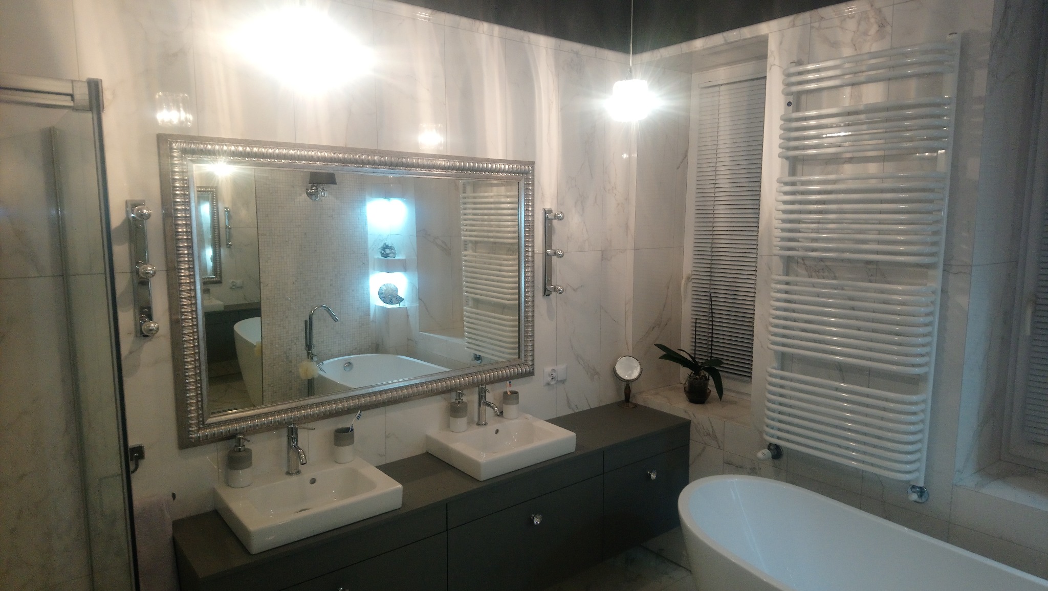 Elegancka łazienka z marmurowymi płytkami, dwoma umywalkami nablatowymi na szarej szafce, dużym lustrem w ozdobnej ramie, grzejnikiem drabinkowym i wolnostojącą wanną.