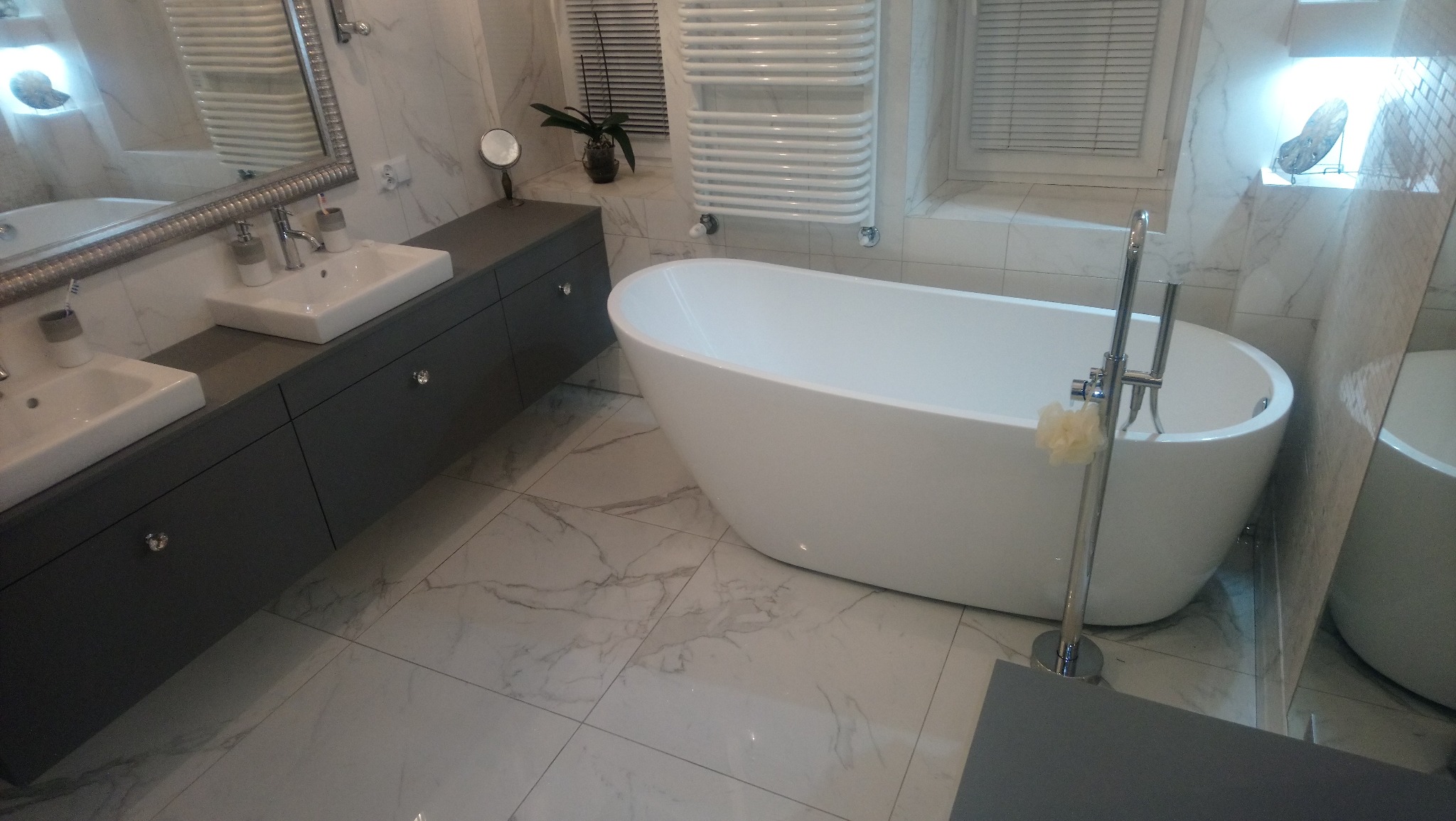 Nowoczesna łazienka z wolnostojącą wanną, dwiema umywalkami nablatowymi na szarym blacie z kryształowymi gałkami, marmurowymi płytkami i białym grzejnikiem drabinkowym