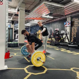 Mężczyzna wykonuje martwy ciąg z obciążeniem, nadzorowany przez drugiego mężczyznę w nowoczesnej siłowni z widocznymi maszynami do ćwiczeń i motywacyjnym napisem na ścianie.