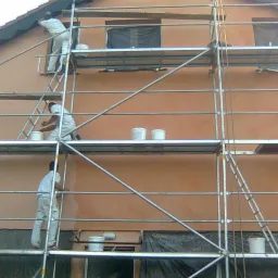 Trzech pracowników w białych kombinezonach na rusztowaniu podczas malowania elewacji budynku na kolor brzoskwiniowy, widoczne wiadra z farbą i zakryte okna.