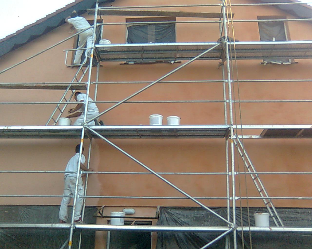 Trzech pracowników w białych kombinezonach na rusztowaniu podczas malowania elewacji budynku na kolor brzoskwiniowy, widoczne wiadra z farbą i zakryte okna.