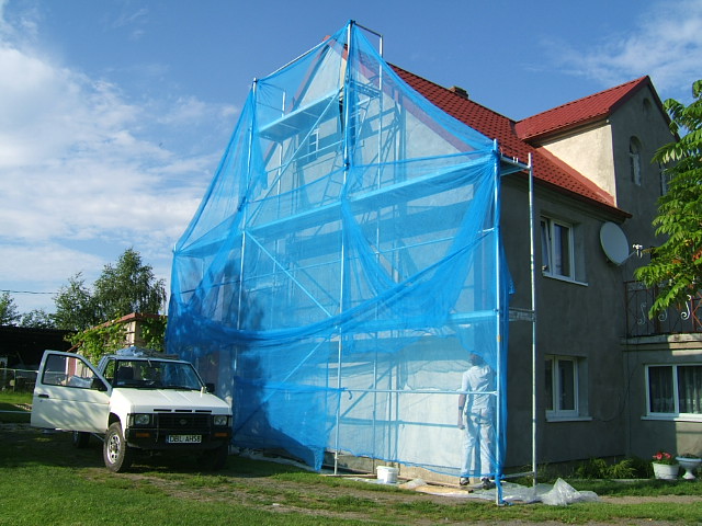 Dom w trakcie ocieplania elewacji, rusztowanie osłonięte niebieską siatką, obok zaparkowany biały samochód dostawczy z otwartymi drzwiami.