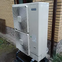 Zewnętrzna jednostka pompy ciepła Hitachi, zamontowana na metalowej konstrukcji przy ścianie budynku z cegły klinkierowej, widoczne ślady deszczu na obudowie.