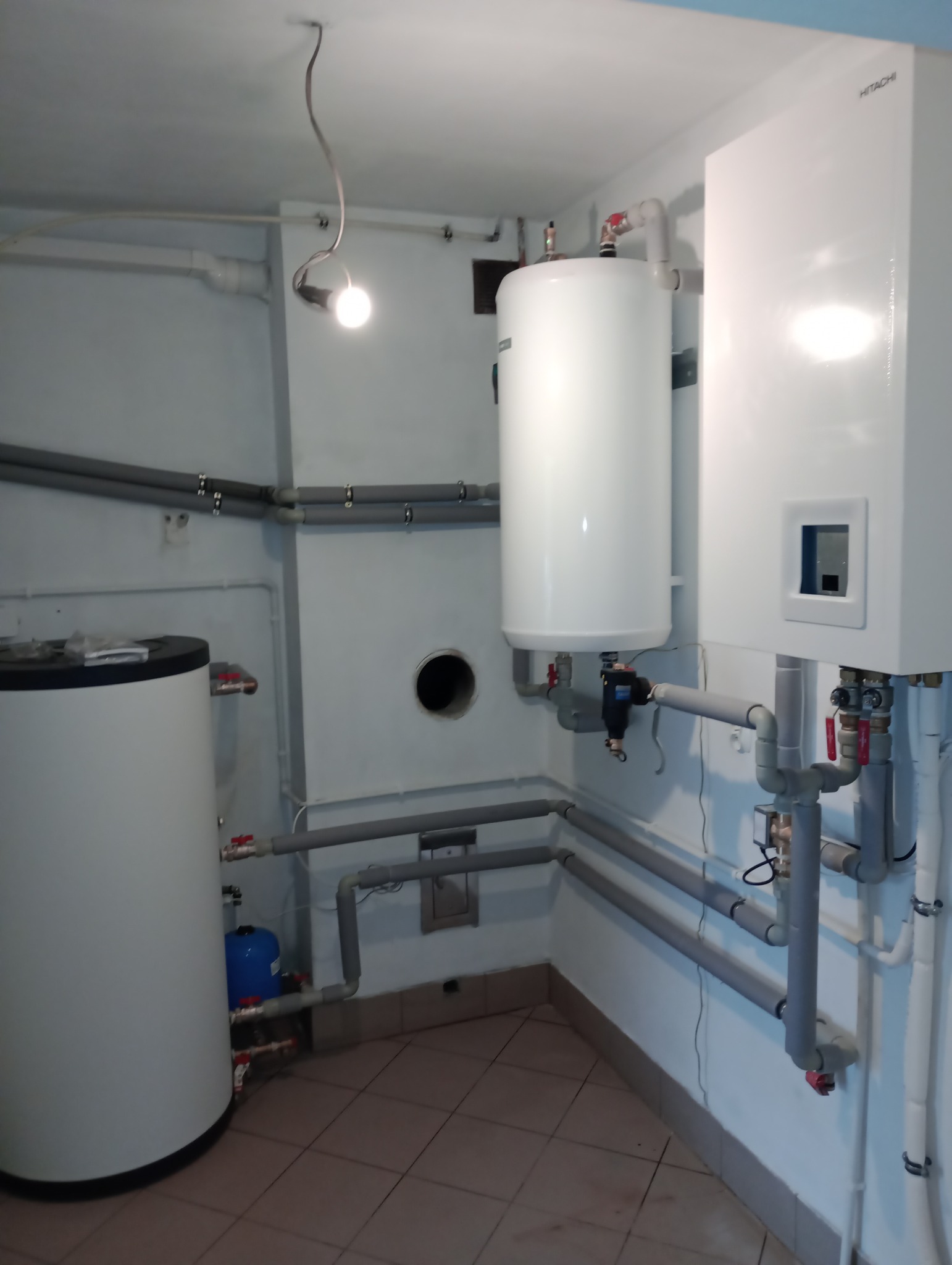 Instalacja grzewcza w kotłowni: biały bojler, zbiornik akumulacyjny i pompa ciepła marki Hitachi, połączone szarymi rurami, oświetlone wiszącą żarówką.