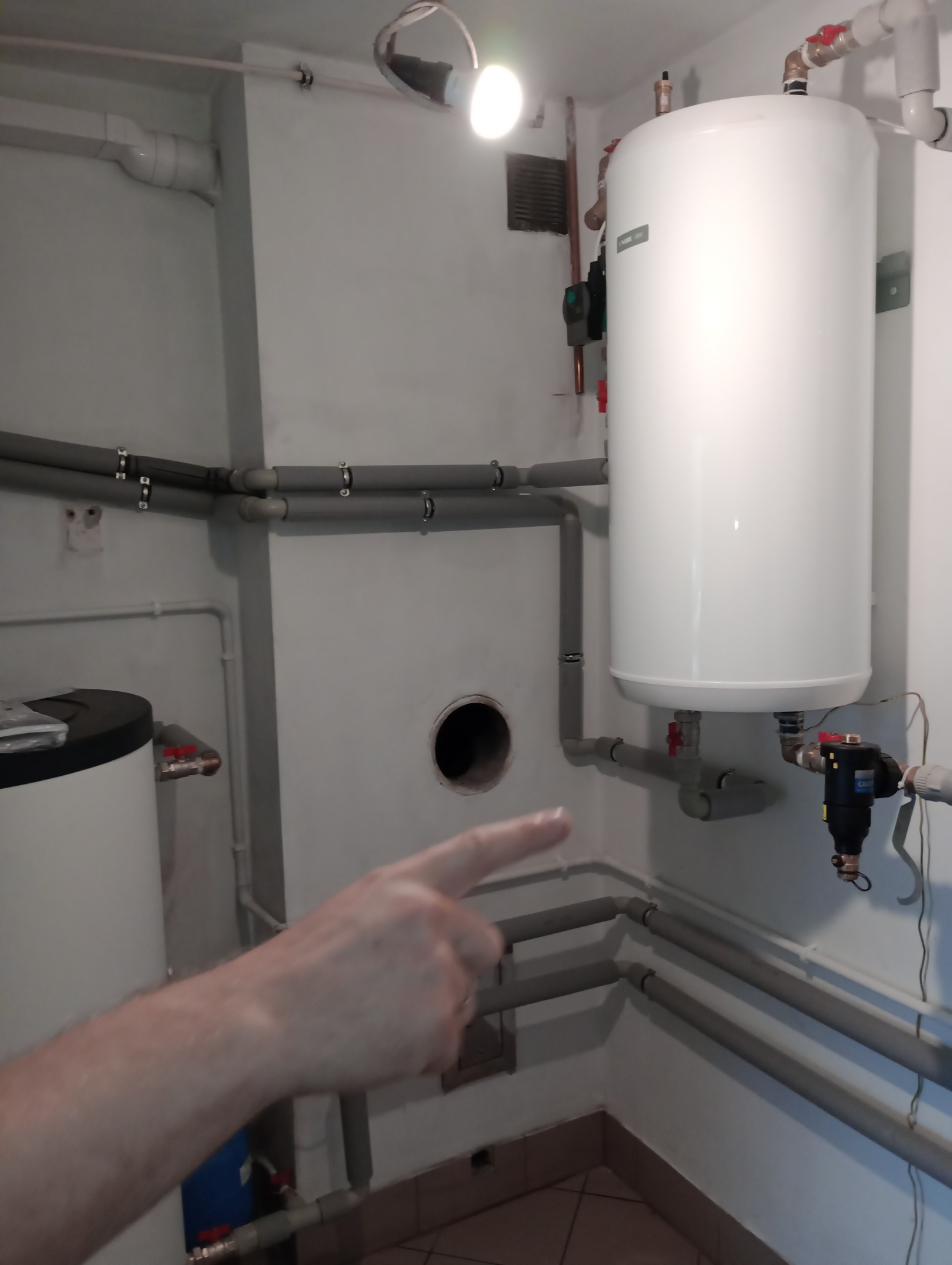 Biały, wiszący zasobnik ciepłej wody Nibe Biawar z widocznymi rurami instalacji grzewczej i ręką wskazującą na otwór w ścianie, oświetlony lampą sufitową.