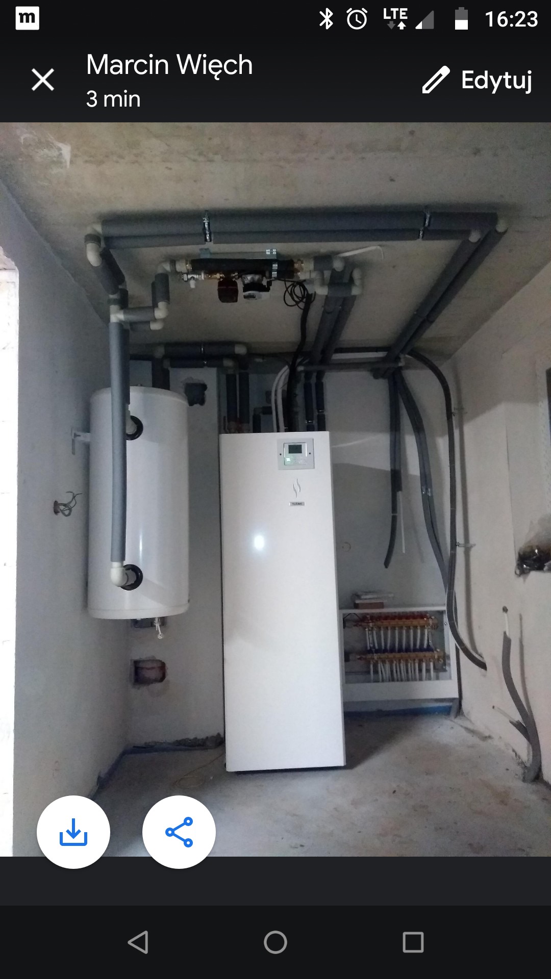 Biały zbiornik i piec grzewczy w surowym pomieszczeniu, z widoczną instalacją rur na suficie i rozdzielaczem podłogowym w tle.