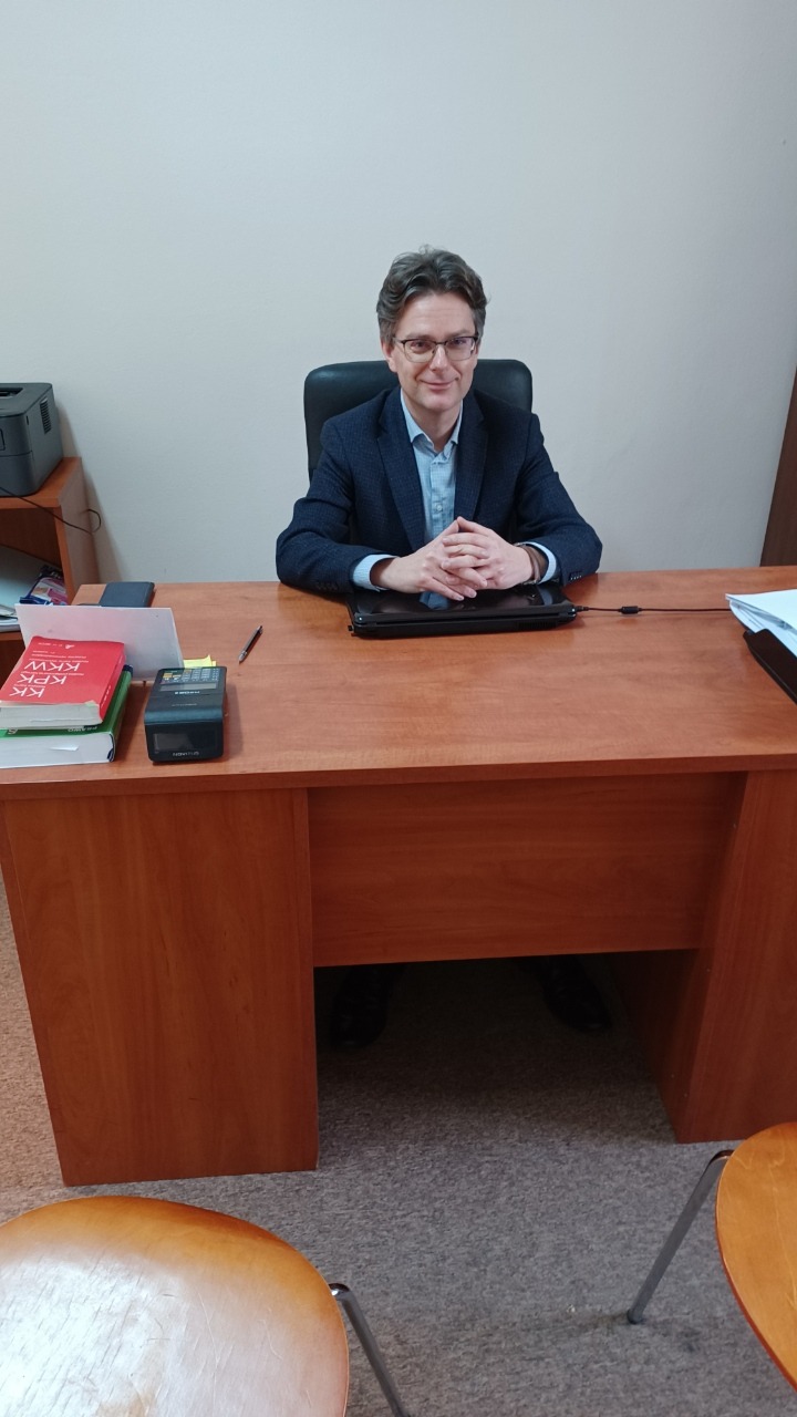 Uśmiechnięty prawnik w okularach siedzi za biurkiem w gabinecie. Na blacie książki prawnicze, notatnik i długopis. Stonowane kolory wnętrza.