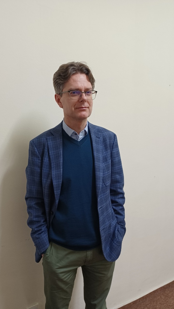 Portret mężczyzny w okularach, ubrany w niebieską marynarkę w kratę i zielone spodnie, stojącego na tle jasnej ściany. Ujęcie od pasa w górę, ręce w kieszeniach.