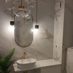 Nowoczesna łazienka z marmurowymi płytkami, owalnym lustrem, wiszącą lampą z szklanymi kulami, białą umywalką nablatową i złotą armaturą.