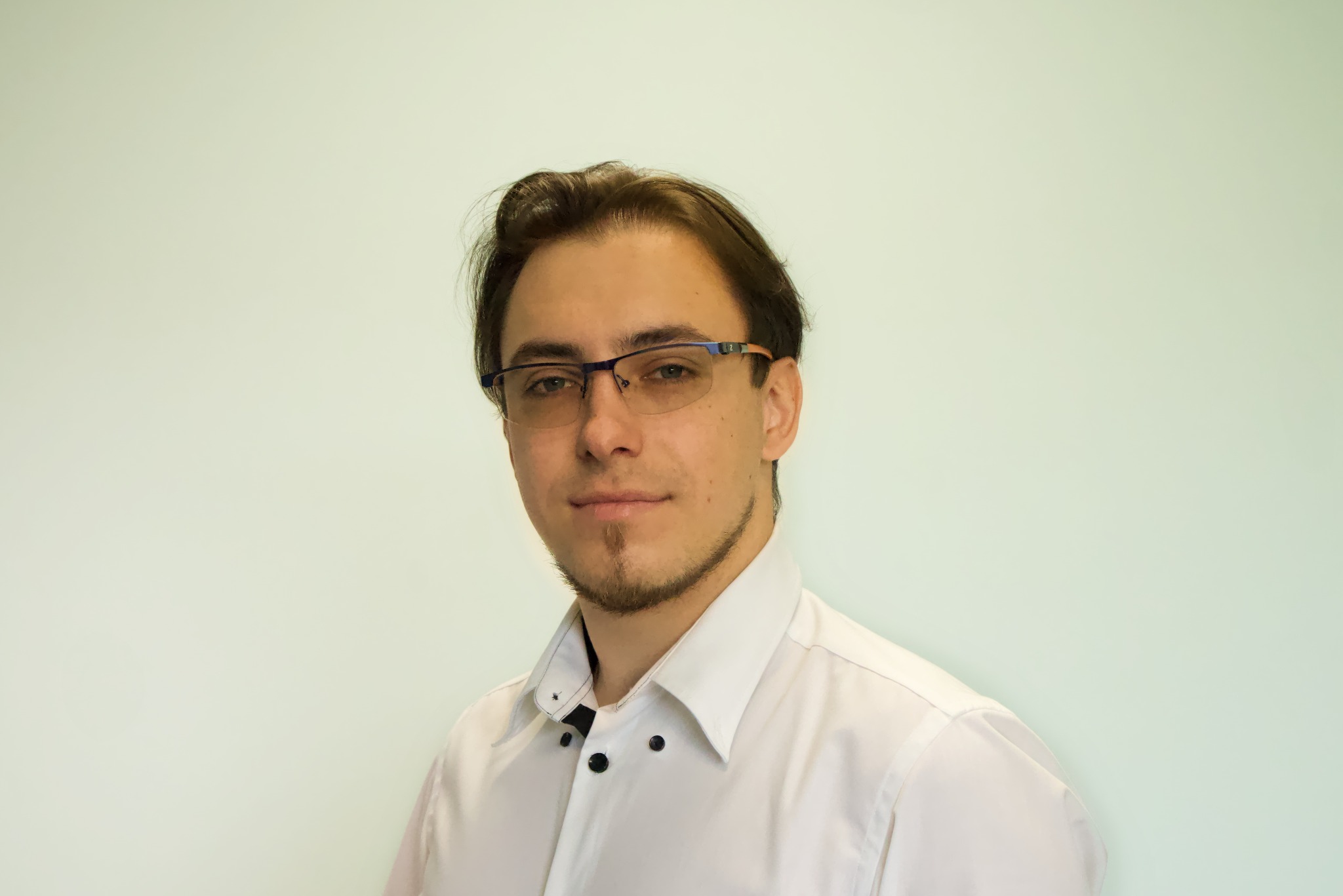 Portret młodego mężczyzny w okularach i białej koszuli na jasnym tle, prawdopodobnie specjalista z Rudy Śląskiej.
