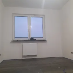 Pusty pokój po szpachlowaniu i malowaniu ścian na biało, podłoga zabezpieczona folią malarską, okno z grzejnikiem pod spodem, widoczne gniazdko elektryczne.