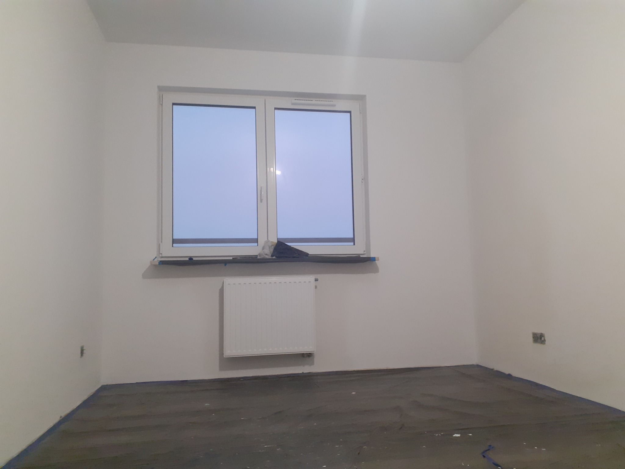Pusty pokój po szpachlowaniu i malowaniu ścian na biało, podłoga zabezpieczona folią malarską, okno z grzejnikiem pod spodem, widoczne gniazdko elektryczne.
