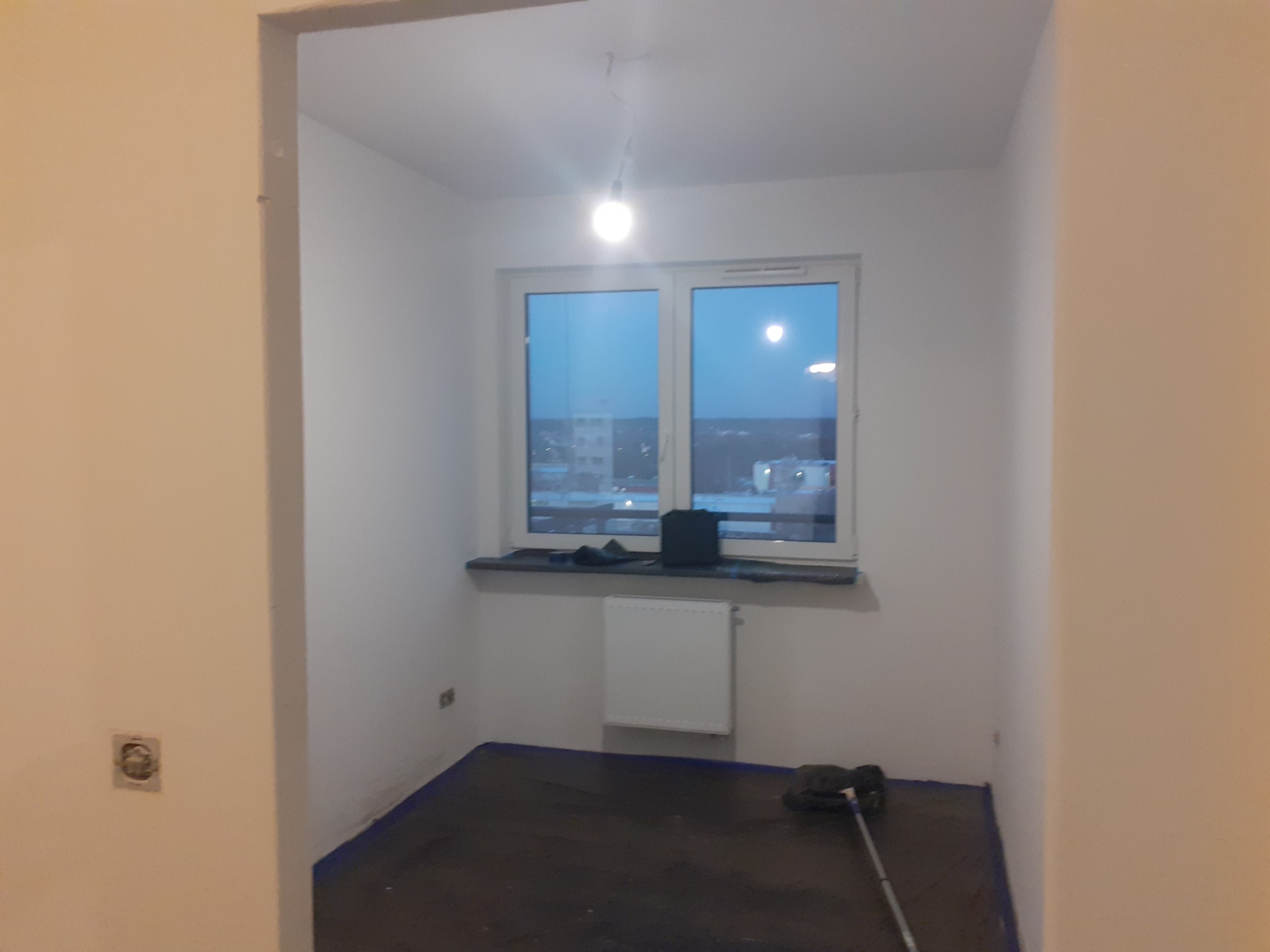 Pokój w trakcie remontu: zabezpieczona folią podłoga, świeżo pomalowane białe ściany, grzejnik, widok z okna na miasto o zmierzchu, lampa na suficie.