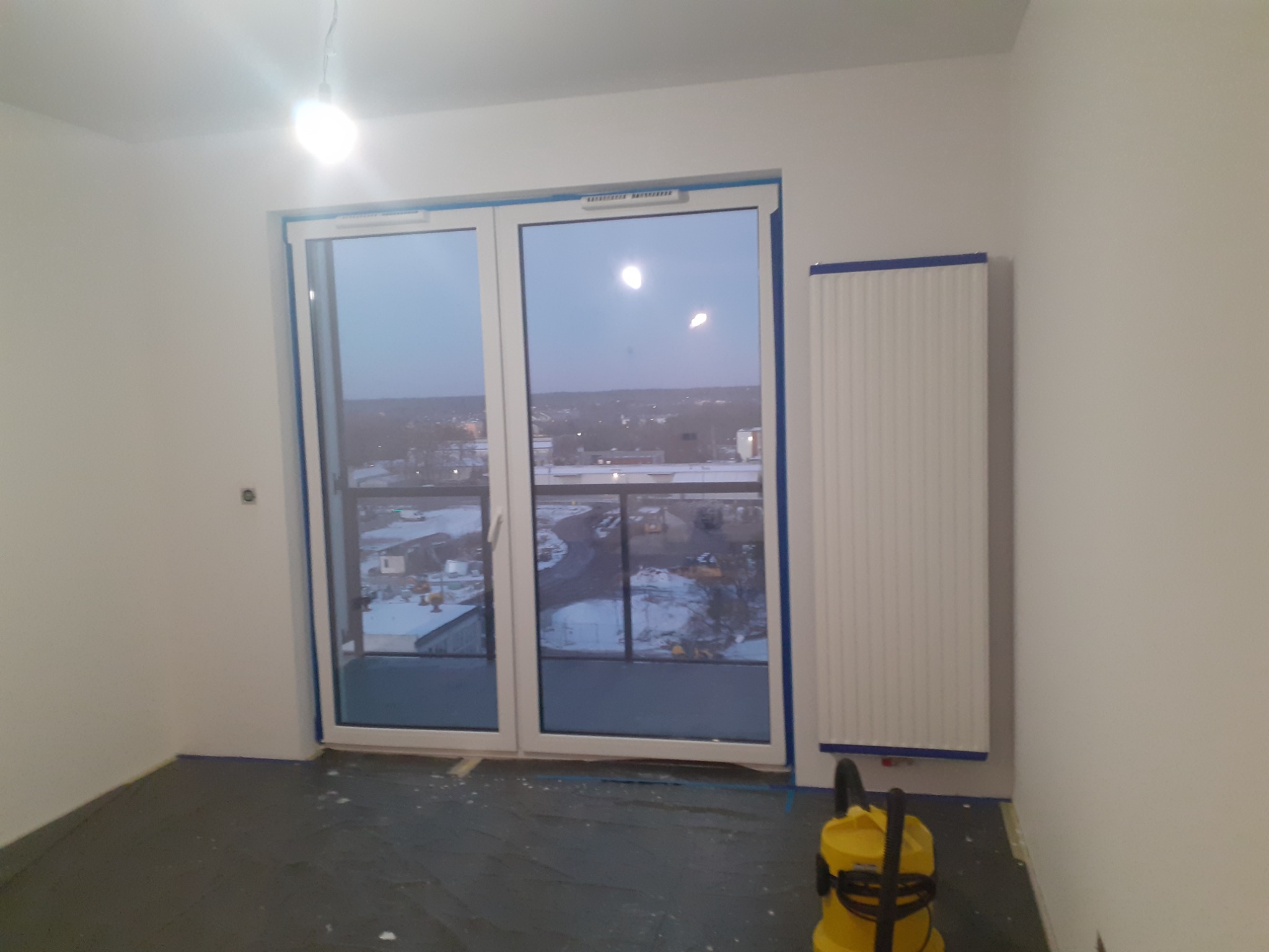 Przygotowane do malowania wnętrze pokoju z zabezpieczoną folią podłogą, grzejnikiem i ramą okienną. Widoczny odkurzacz przemysłowy.