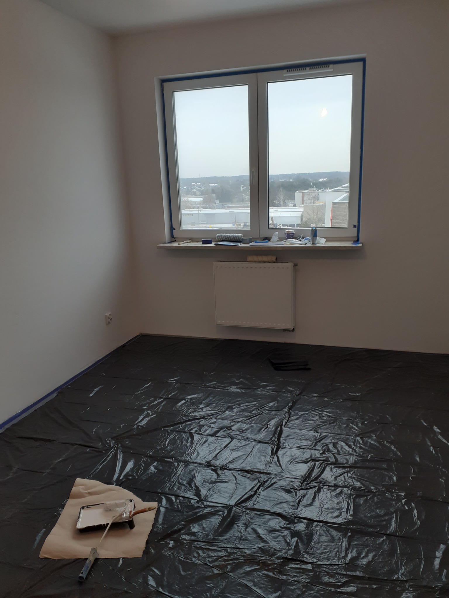 Przygotowane do malowania wnętrze pokoju: podłoga zabezpieczona czarną folią malarską, okno i listwy przypodłogowe oklejone taśmą malarską, widoczny wałek i kuweta z jasną farbą.