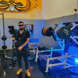 Kamerzysta w okularach przeciwsłonecznych, ubrany w uprząż stabilizującą kamerę, stoi w siłowni z urządzeniami do ćwiczeń siłowych.
