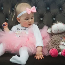 Dziewczynka w białych rajstopach i różowej tiulowej spódniczce siedzi na ciemnobrązowej kanapie obok pluszowej owieczki.