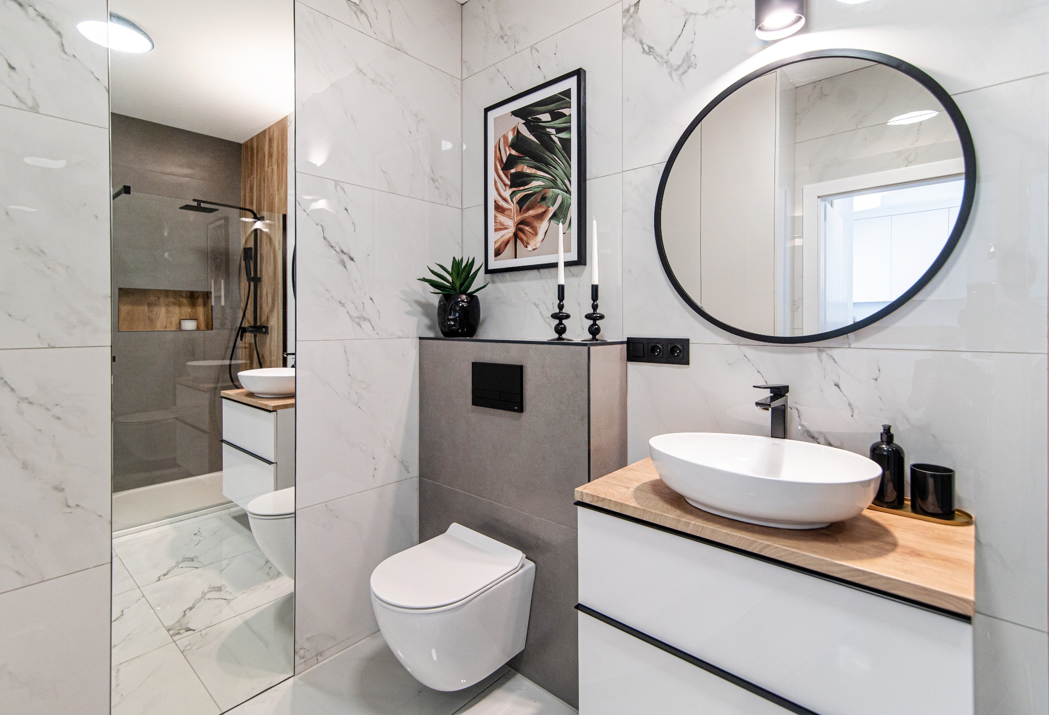 Nowoczesna łazienka z marmurowymi płytkami, owalnym lustrem w czarnej ramie, wiszącą toaletą i umywalką nablatową na drewnianym blacie, widok na przeszkloną kabinę prysznicową.