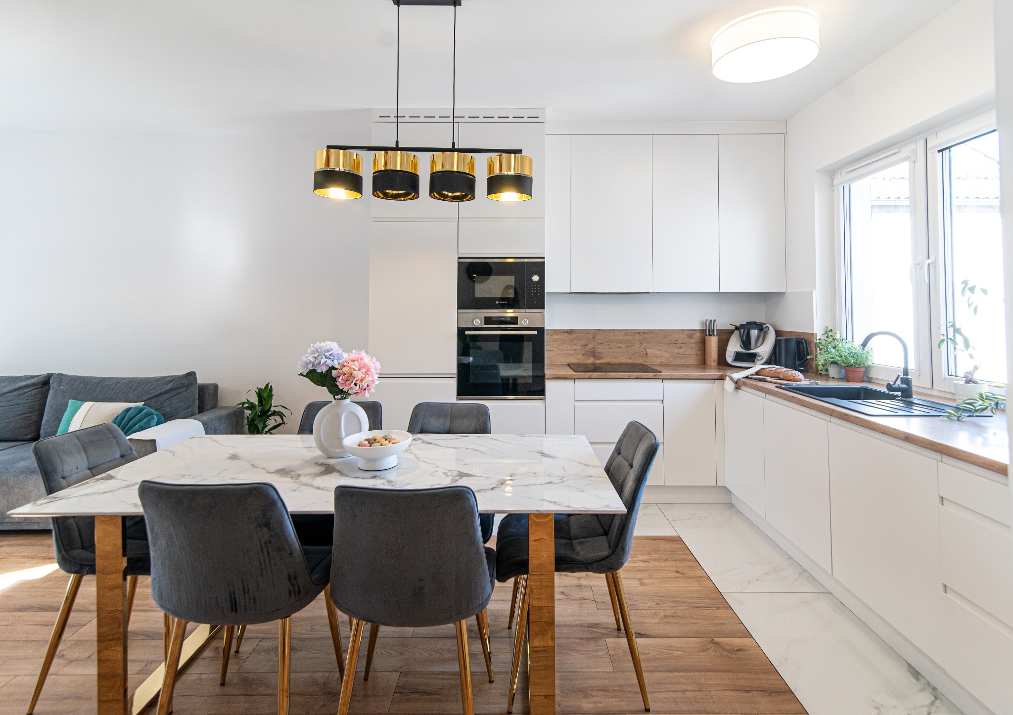 Jasne wnętrze jadalni z marmurowym stołem, szarymi krzesłami na złotych nogach, nowoczesną lampą i widokiem na kuchnię z białymi szafkami i drewnianym blatem.