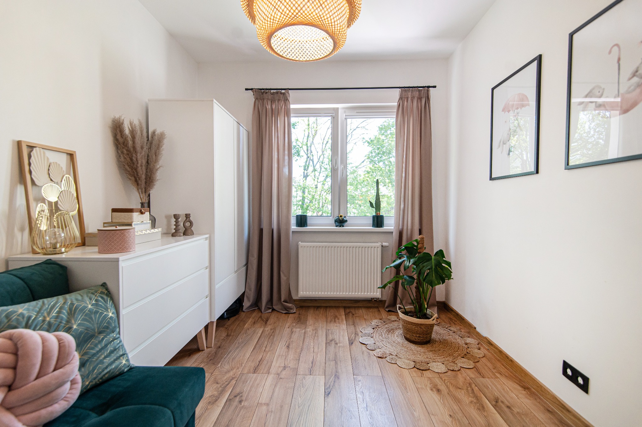 Jasne wnętrze pokoju z drewnianą podłogą, białymi ścianami, komodą z dekoracjami, zieloną kanapą i roślinami w doniczkach.