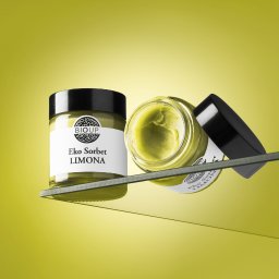 Paweł Retkowski Studio - Dwa słoiczki z żółtym kremem Eko Sorbet Limona marki BIOUP, jeden stojący, drugi przechylony z otwartym wieczkiem, na szklanej półce na tle jednolitego, żółtego tła.